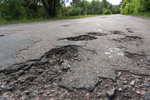 Rough road with large potholes and greenery around.